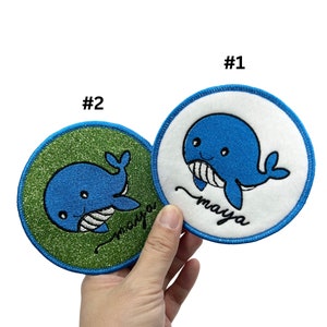 May include: Two embroidered patches featuring a blue whale with a white belly and the name "maya" written in cursive. One patch has a green glitter background and the other has a white background.