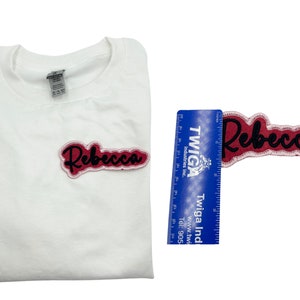May include: A white t-shirt with a pink and black iron-on patch that says "Rebecca".