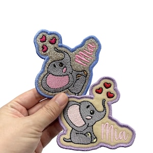 May include: Two embroidered iron-on patches featuring a gray elephant with pink ears and a pink heart. The top patch has a blue border and the text "Mia" in pink. The bottom patch has a purple border and the text "Mia" in white.