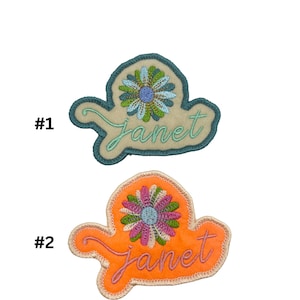 May include: Two embroidered iron-on patches featuring a flower design and the name "Janet" in cursive. The first patch is on a beige background with teal stitching. The second patch is on an orange background with pink stitching.