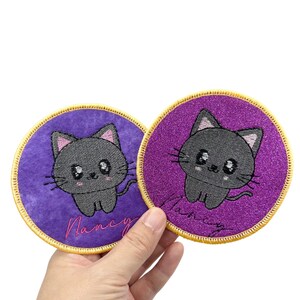 May include: Two purple embroidered iron-on patches featuring a black cat design. One patch has a glittery background and the other has a smooth background. Both patches have the name "Nancy" embroidered on them.