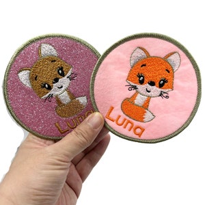 May include: Two round pink and peach fabric patches with embroidered fox faces and the name "Luna" in orange thread.