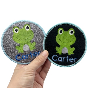 May include: Two round iron-on patches featuring a green frog design and the name "Carter" embroidered in blue. One patch has a silver glitter background and the other has a black background.