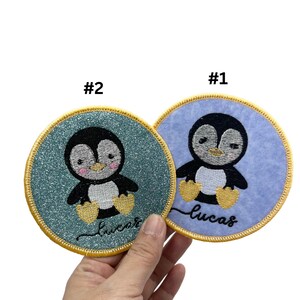 May include: Two round embroidered patches featuring a penguin design. The penguin is black and white with a yellow beak and feet. The patches are edged with a gold border. The text "Lucas" is embroidered on each patch. One patch has a blue background and the other has a green background.