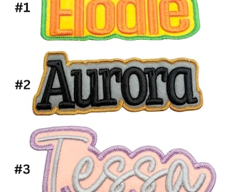 May include: Three embroidered iron-on patches with names: Elodie in yellow and green, Aurora in gray and black, and Tessa in pink and purple.