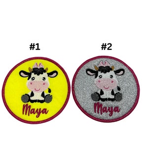 May include: Two embroidered iron-on patches featuring a cartoon cow with black and white spots. The cow is sitting down with its legs crossed. The patches are round and have a pink border. The patches are on a white background. The text "Maya" is embroidered on the patches in pink. The patch on the left is yellow and the patch on the right is silver.