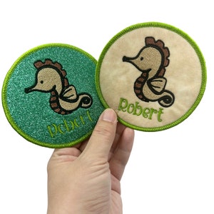 Peut inclure: Deux patchs ronds brodés avec un motif de cheval de mer brun. Un patch est vert avec des paillettes et dit "Pebble" et l'autre est beige avec le nom "Robert".