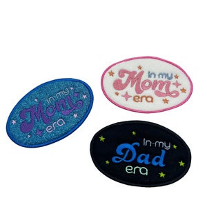 Peut inclure: Trois patchs brodés thermocollants avec les mots "In my Mom era", "In my Dad era" et "In my Mom era" en écriture cursive. Les patchs sont de forme ovale et ont un fond blanc, noir et bleu.