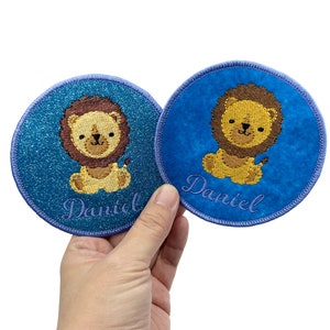 May include: Two blue and turquoise round iron-on patches with a lion design and the name "Daniel" embroidered in purple thread.