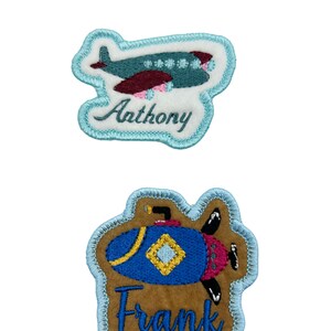 May include: Two embroidered iron-on patches with blue borders. The top patch features a blue airplane with a pink and maroon design and the name "Anthony" embroidered in blue. The bottom patch features a blue submarine with a yellow and maroon design and the name "Frank" embroidered in brown.