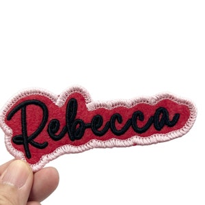 May include: Red felt patch with a pink outline and the name "Rebecca" embroidered in black.