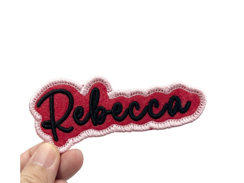 May include: Red felt patch with a pink outline and the name "Rebecca" embroidered in black.
