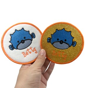 May include: Two circular patches with a blue pufferfish design and the name "Betty" in orange. One has a white background with an orange border, the other a gold glitter background with an orange border.