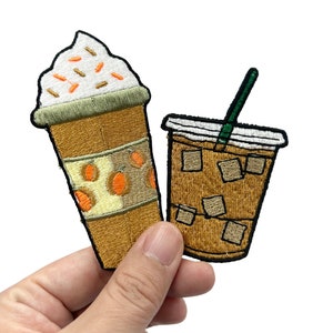 May include: Embroidered patches of coffee drinks. One shows a coffee with cream and sprinkles, the other an iced coffee with a green straw. The patches are brown and tan in colour.