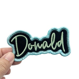 May include: A blue and black embroidered patch with the word "Donald" in white thread.