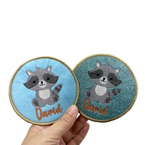 May include: Two round embroidered patches featuring a raccoon design. The patches are blue and teal with gold trim. The name "David" is embroidered on each patch in orange thread.