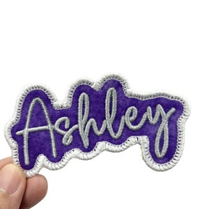 May include: Purple and silver embroidered patch with the name "Ashley" in a cursive font.