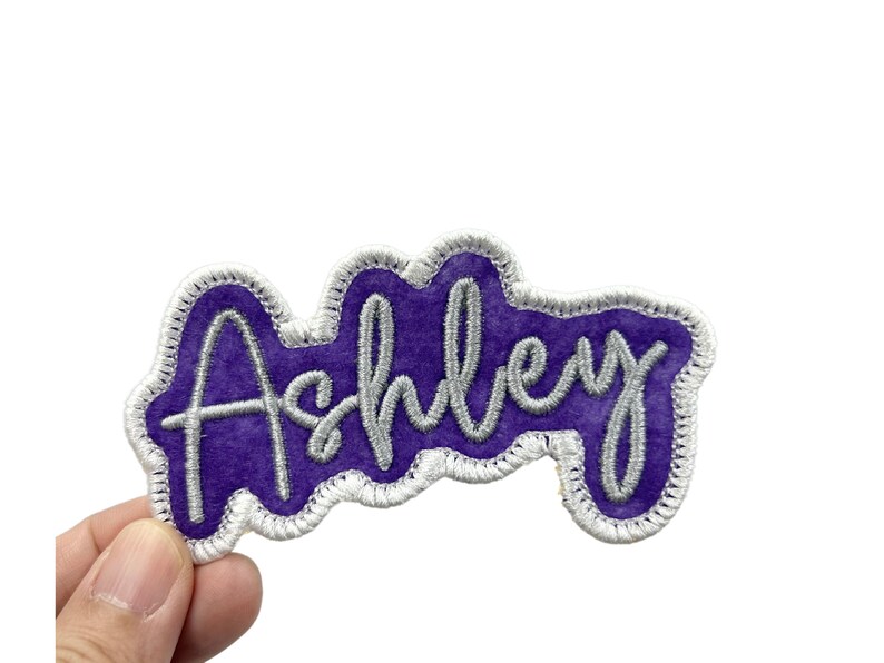 May include: Purple and silver embroidered patch with the name "Ashley" in a cursive font.