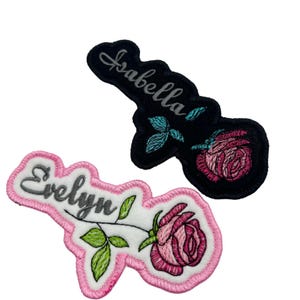 May include: Two embroidered iron-on patches featuring pink roses and names. The top patch is black with the name "Isabella" in silver thread. The bottom patch is pink with the name "Evelyn" in silver thread.