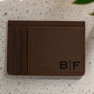 May include: A brown leather card holder with the initials "BF" engraved on the front. The wallet has multiple card slots and is made of a smooth, textured leather. The card holder is rectangular and has a stitched border.