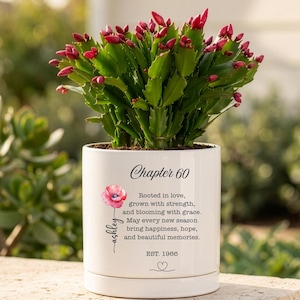 May include: A white ceramic planter containing a vibrant Christmas cactus with green leaves and pink buds. The pot features the text Chapter 60, a pink poppy illustration, the name Ashley, and the quote Rooted in love, grown with strength.
