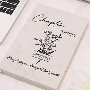 May include: A white marble-patterned notebook with the words "Chapter Thirty" and "Christina" printed on the cover. A floral illustration is centered below the text. The phrase "Every Chapter Brings New Growth" is printed at the bottom.