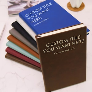 May include: A stack of notebooks in various colors, including blue, brown, and gray. Each notebook has the text "CUSTOM TITLE YOU WANT HERE" and "Custom Subtitle" printed on the cover. The notebooks are arranged on a white surface.