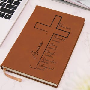 May include: A tan leather-like journal featuring a black printed cross design. The text reads: Anna, You are Beautiful, Strong, Enough, Never alone, Always loved, along with Bible verse references. It includes a matching tan ribbon bookmark.