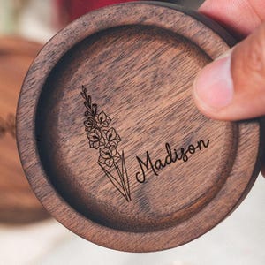 May include: A round, wooden coaster with a floral design and the name "Madison" engraved on it. The coaster is made of dark brown wood and has a smooth finish. The floral design is a delicate line drawing.