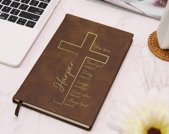 May include: A brown leather journal with gold lettering. The cover features a cross design and the name "Harper". Text includes "You are Beautiful, Strong, Enough, Never alone, Always loved". A ribbon bookmark is visible.