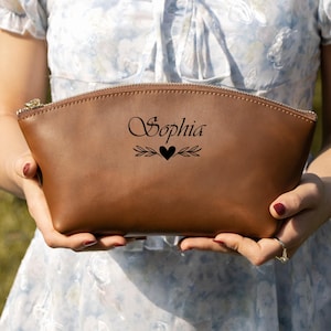 K&ouml;nnte beinhalten: Eine braune Kosmetiktasche aus Leder mit goldenem Rei&szlig;verschluss, gehalten von einer Person. Auf der Tasche steht der Name "Sophia" in eleganter Schrift, zusammen mit einem Herz- und Bl&auml;ttermotiv. Die Tasche hat eine warme braune Farbe.