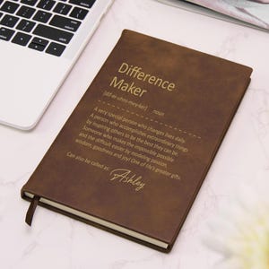 May include: A brown notebook with a gold-printed definition of the term Difference Maker, including the phonetic spelling and a descriptive paragraph. The text also features the name Ashley in a script font at the bottom, with a ribbon bookmark.