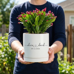 May include: A white cylindrical ceramic planter containing a green Christmas cactus with numerous pink flower buds. The front of the planter features the black printed text "olivia" followed by a heart symbol, "david", and the date "09.12.2026".