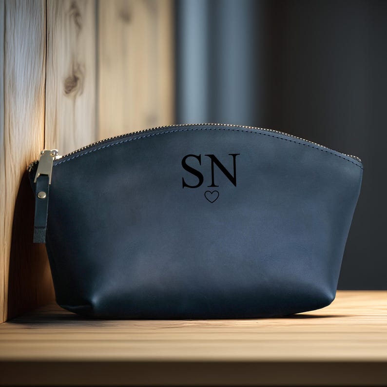 K&ouml;nnte beinhalten: Eine marineblaue Rei&szlig;verschlusstasche mit goldfarbener Hardware. Die Tasche tr&auml;gt die Initialen "SN" mit einem Herzsymbol darunter. Sie besteht aus glattem Material und liegt auf einer Holzoberfl&auml;che.