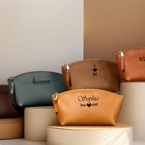 K&ouml;nnte beinhalten: Eine Sammlung personalisierter Kosmetiktaschen in verschiedenen Farben, darunter Braun, Blaugr&uuml;n und Beige. Jede Tasche hat einen goldenen Rei&szlig;verschluss und ist mit einem Namen oder Initialen in Schwarz beschriftet. Die Taschen werden auf zylindrischen Sockeln pr&auml;sentiert.