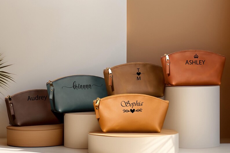 K&ouml;nnte beinhalten: Eine Sammlung personalisierter Kosmetiktaschen in verschiedenen Farben, darunter Braun, Blaugr&uuml;n und Beige. Jede Tasche hat einen goldenen Rei&szlig;verschluss und ist mit einem Namen oder Initialen in Schwarz beschriftet. Die Taschen werden auf zylindrischen Sockeln pr&auml;sentiert.