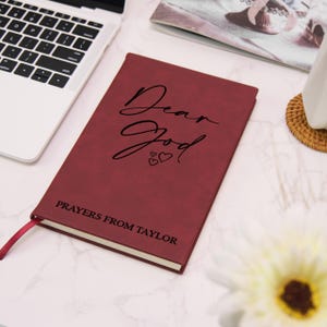 Puede incluir: Un diario burdeos con las palabras "Dear God" en escritura elegante, acompa&ntilde;ado de dos corazones. La parte inferior del diario dice "Prayers From Taylor". Una cinta roja como marcador es visible. El diario est&aacute; sobre una superficie de m&aacute;rmol blanco.