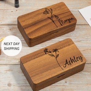 May include: Two rectangular wooden boxes with engraved floral designs and names. One box is labeled "Morgan," and the other is labeled "Ashley." The boxes are made of a warm-toned wood and have a simple, elegant design.