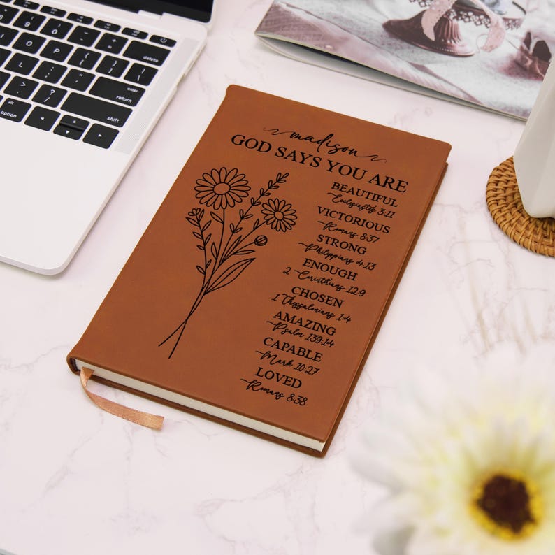 Puede incluir: Un diario de cuero marr&oacute;n con el texto "madison GOD SAYS YOU ARE" y un dise&ntilde;o floral. El diario incluye una cinta de marcador y una lista de palabras y vers&iacute;culos b&iacute;blicos inspiradores.