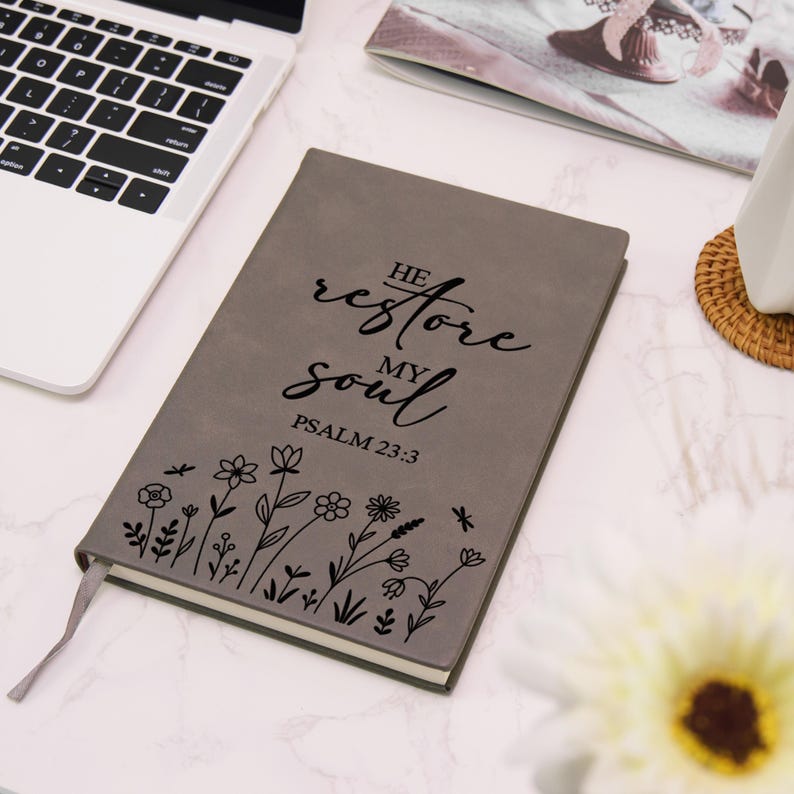 Puede incluir: Diario gris con el texto "He restore my soul Psalm 23:3" en escritura negra. El diario presenta un dise&ntilde;o floral y una cinta de marcador gris. Est&aacute; sobre una superficie blanca cerca de un port&aacute;til.