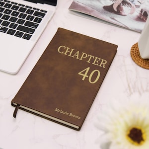 May include: A brown leather journal with the words "CHAPTER 40" in gold lettering. The name "Melanie Brown" is printed at the bottom. A ribbon bookmark is visible. The journal is on a white surface.