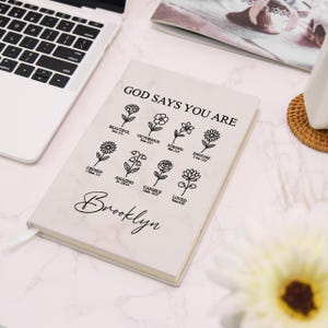 Puede incluir: Un cuaderno blanco con dise&ntilde;o de m&aacute;rmol con el texto "GOD SAYS YOU ARE" e ilustraciones florales. Debajo est&aacute; el nombre "Brooklyn" en cursiva. El cuaderno est&aacute; abierto, revelando p&aacute;ginas rayadas y una cinta de marcador.