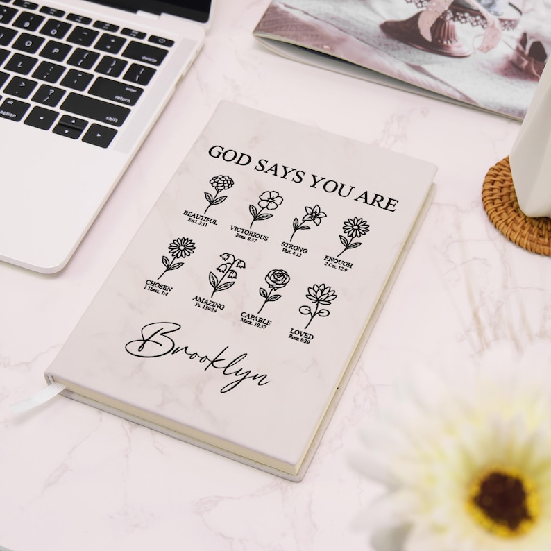 Puede incluir: Un cuaderno blanco con dise&ntilde;o de m&aacute;rmol con el texto "GOD SAYS YOU ARE" e ilustraciones florales. Debajo est&aacute; el nombre "Brooklyn" en cursiva. El cuaderno est&aacute; abierto, revelando p&aacute;ginas rayadas y una cinta de marcador.
