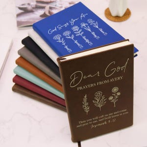 Puede incluir: Una pila de diarios en varios colores, incluyendo azul, marr&oacute;n y gris. El diario frontal es marr&oacute;n con las palabras "Dear God" y "Prayers From Avery" impresas en la portada. El diario azul tiene el texto "God Says You Are" e ilustraciones florales.
