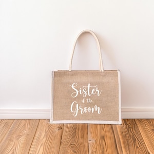 Pode incluir: Uma bolsa de compras de juta marrom com al&ccedil;as brancas e o texto "Sister of the Groom" impresso na frente.