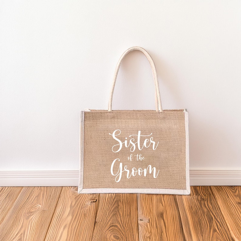 Pode incluir: Uma bolsa de compras de juta marrom com al&ccedil;as brancas e o texto "Sister of the Groom" impresso na frente.