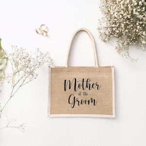 Pode incluir: Uma bolsa de compras de juta marrom com acabamento branco e al&ccedil;as. A bolsa tem o texto "Mother of the Groom" impresso em preto.
