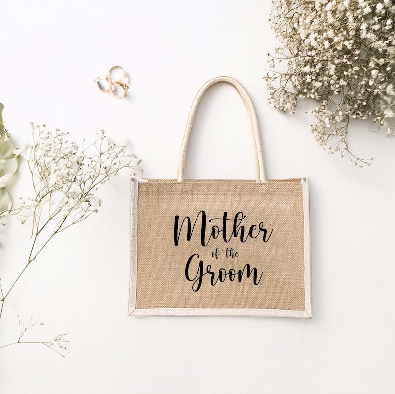 Pode incluir: Uma bolsa de compras de juta marrom com acabamento branco e al&ccedil;as. A bolsa tem o texto "Mother of the Groom" impresso em preto.