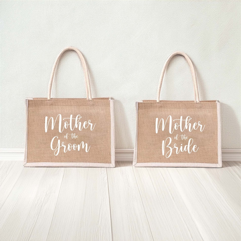Pode incluir: Duas sacolas de compras de juta marrom com al&ccedil;as brancas e texto branco. A sacola &agrave; esquerda diz "M&atilde;e do noivo" e a sacola &agrave; direita diz "M&atilde;e da noiva".