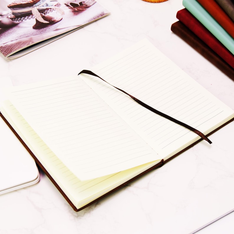 Puede incluir: Un cuaderno abierto con p&aacute;ginas rayadas de color crema y una cubierta de cuero marr&oacute;n. Una cinta marr&oacute;n como marcador de libros cruza las p&aacute;ginas abiertas. Otros cuadernos de varios colores est&aacute;n apilados en el fondo, sobre una superficie blanca.